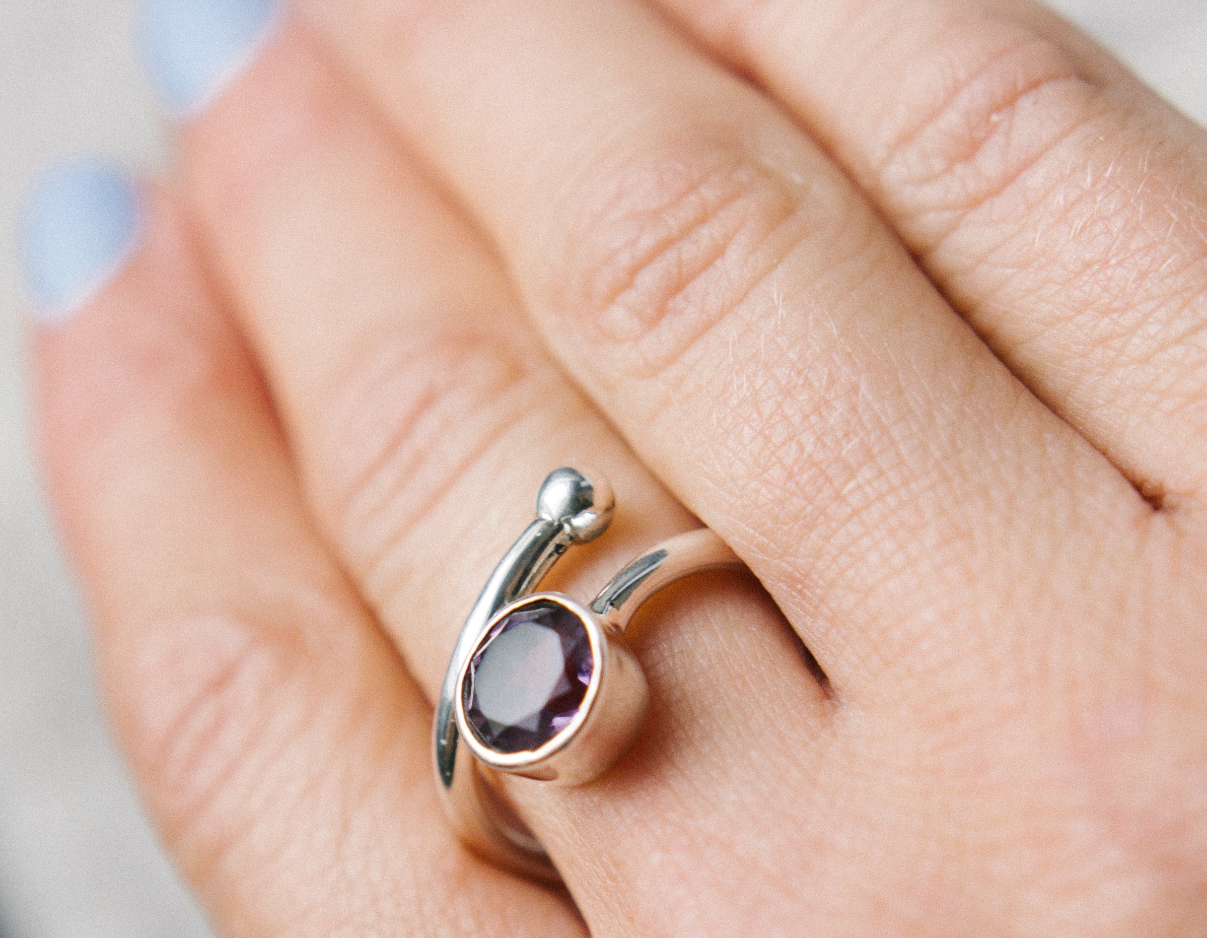 Close-up of a hand with light blue painted nails wearing the Honeysuckle Ring in Silver Gold & gemstones by Gallardo & Blaine Designs. The unique silver design features an open-ended band with a shiny round purple gemstone in a minimalist bezel setting on
