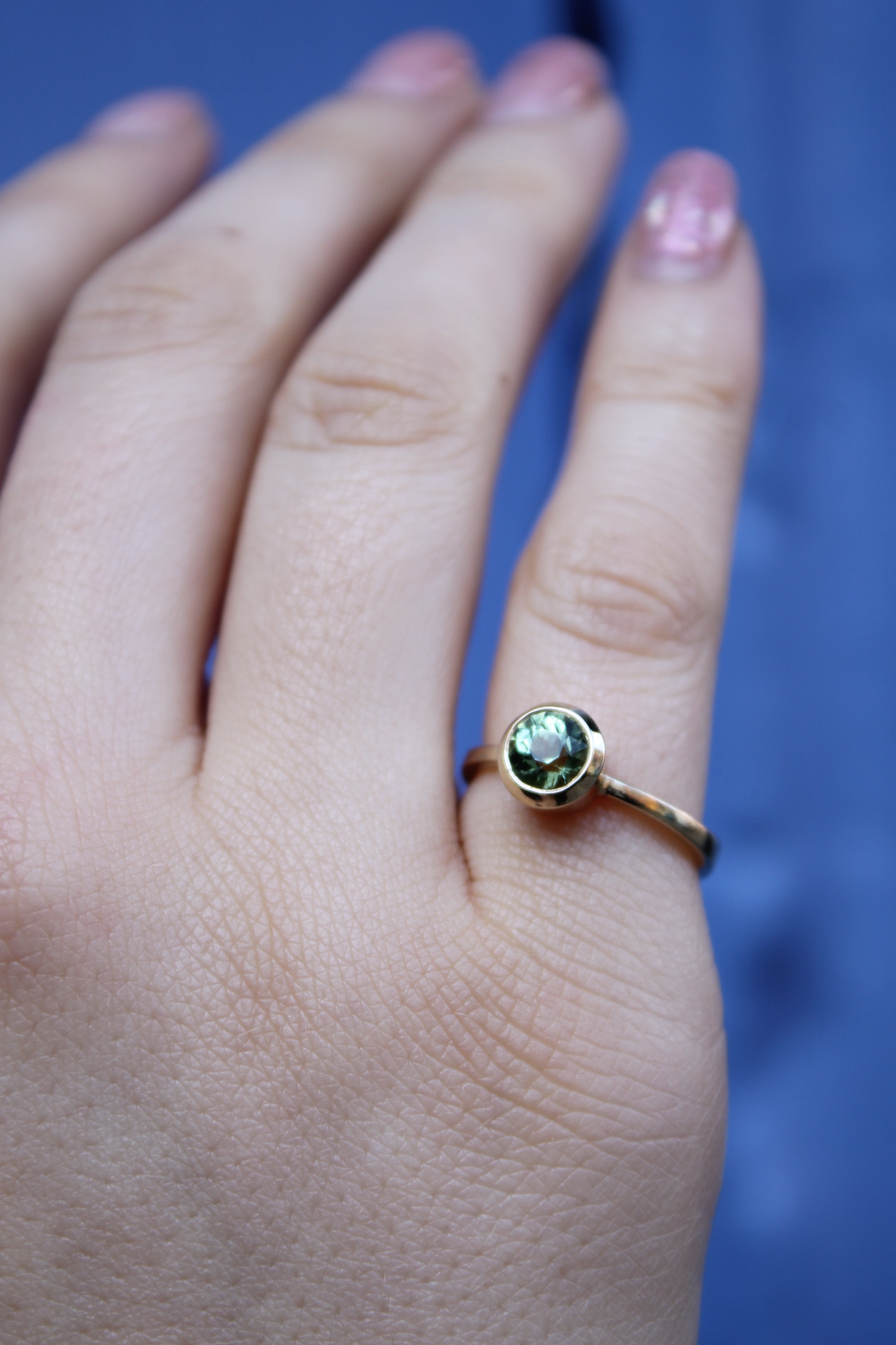 A hand with natural nails wears the Madeleine Fine Jewellery Viridian Dawn 9ct yellow gold ring, set with a small round green beryl, against a blurred blue background.