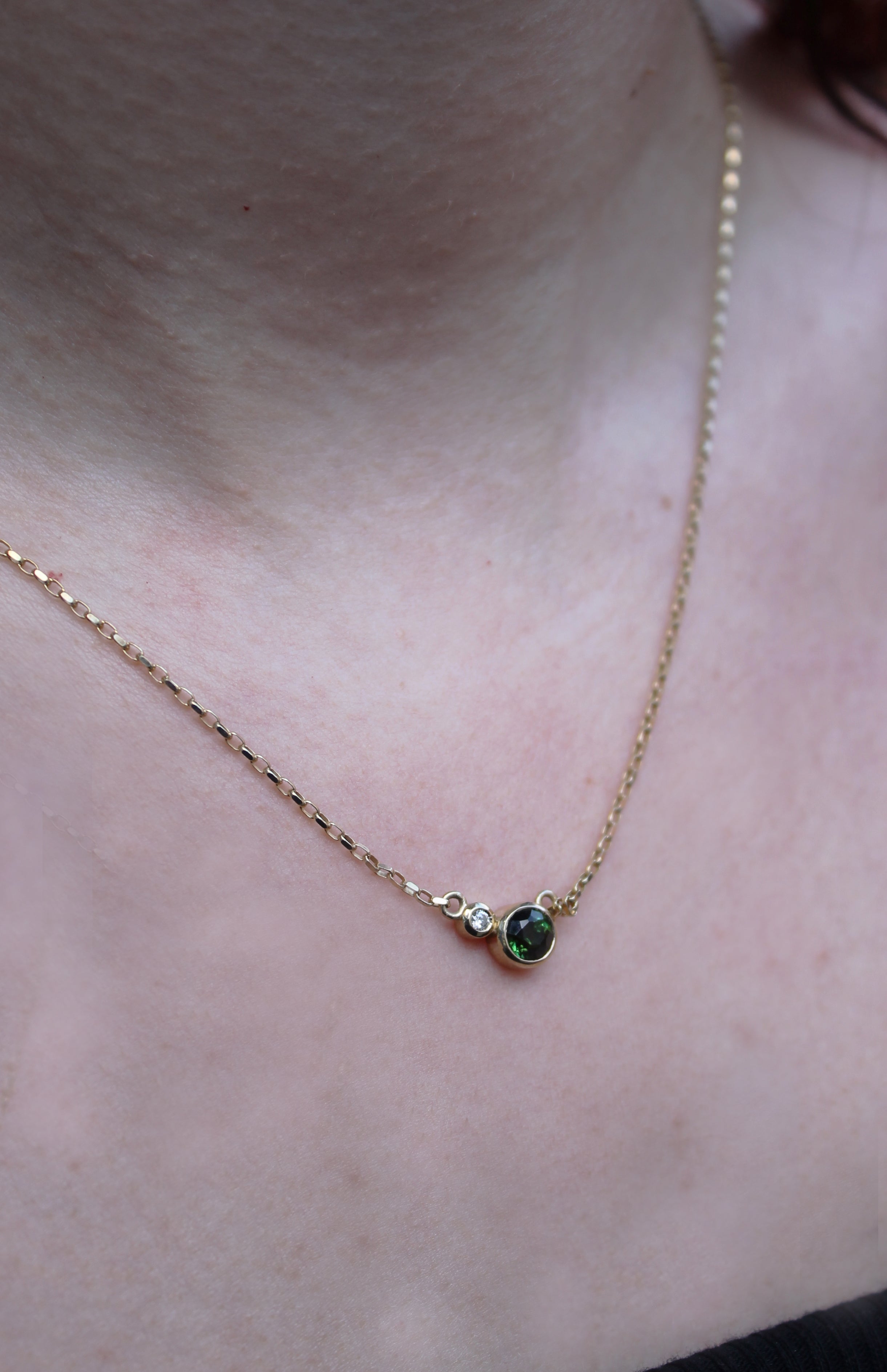 A close-up of a person in a black top wearing the Madeleine Fine Jewellery Verdant Embrace 9ct Yellow Gold Necklace, featuring a lab-grown diamond pendant and a larger round green tourmaline pendant.