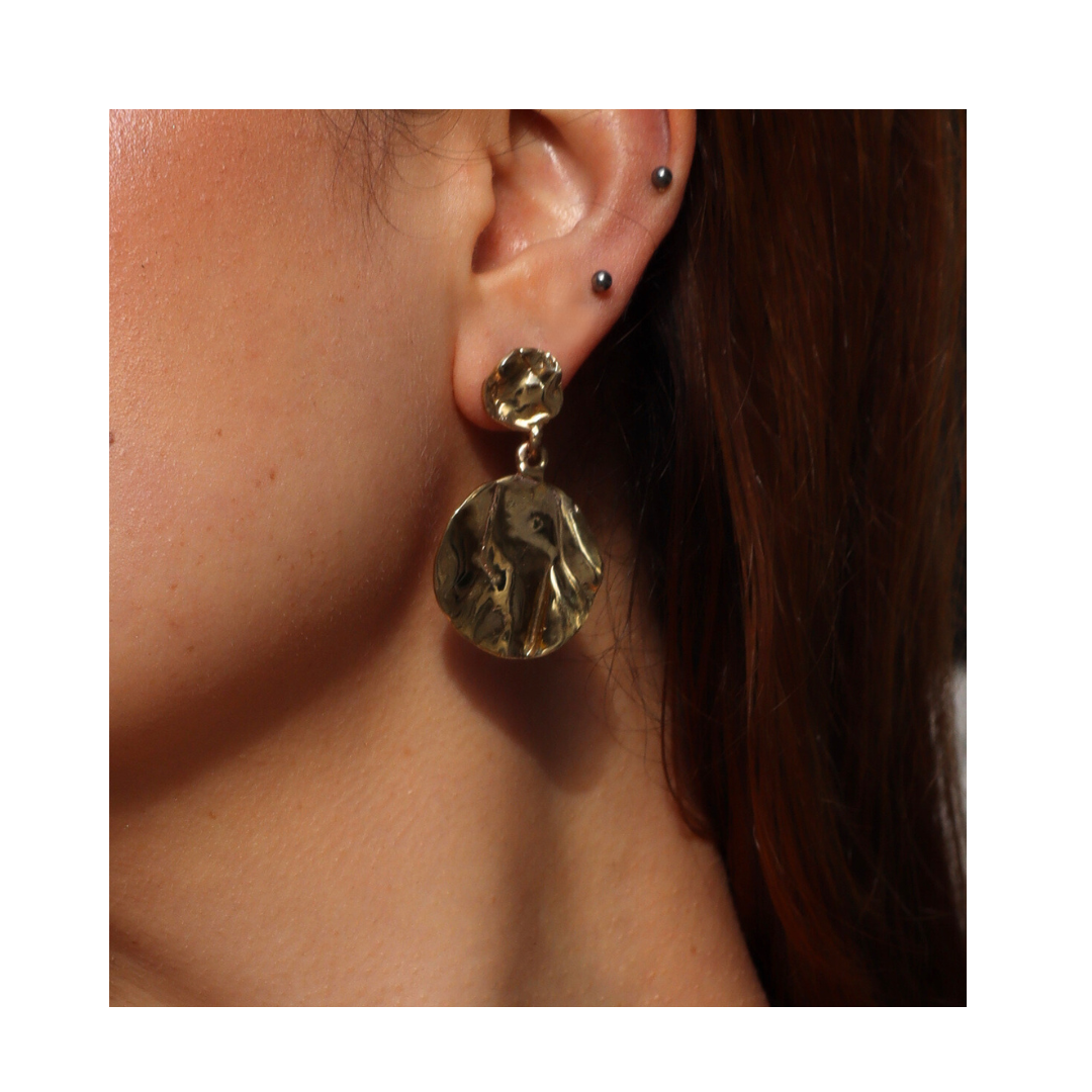 Close-up of a person’s ear with three small black piercings and a large, gold, textured Tide Drop Earring by Madeleine Sculptural Jewellery—crafted from sustainable materials—in the lobe. Brown hair frames the side of the head.