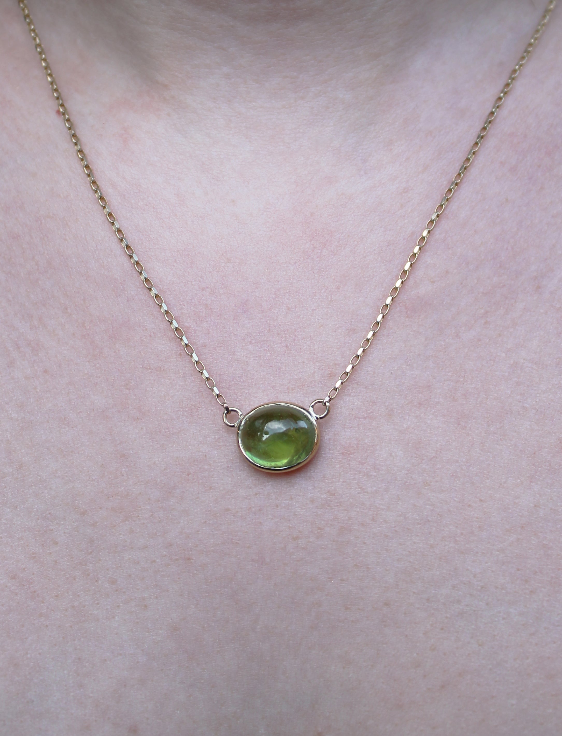 A close-up of the Whispering Moss 9ct Yellow Gold Necklace by Madeleine Fine Jewellery, showcasing a round green peridot cabochon pendant resting on a person's bare neck and upper chest.