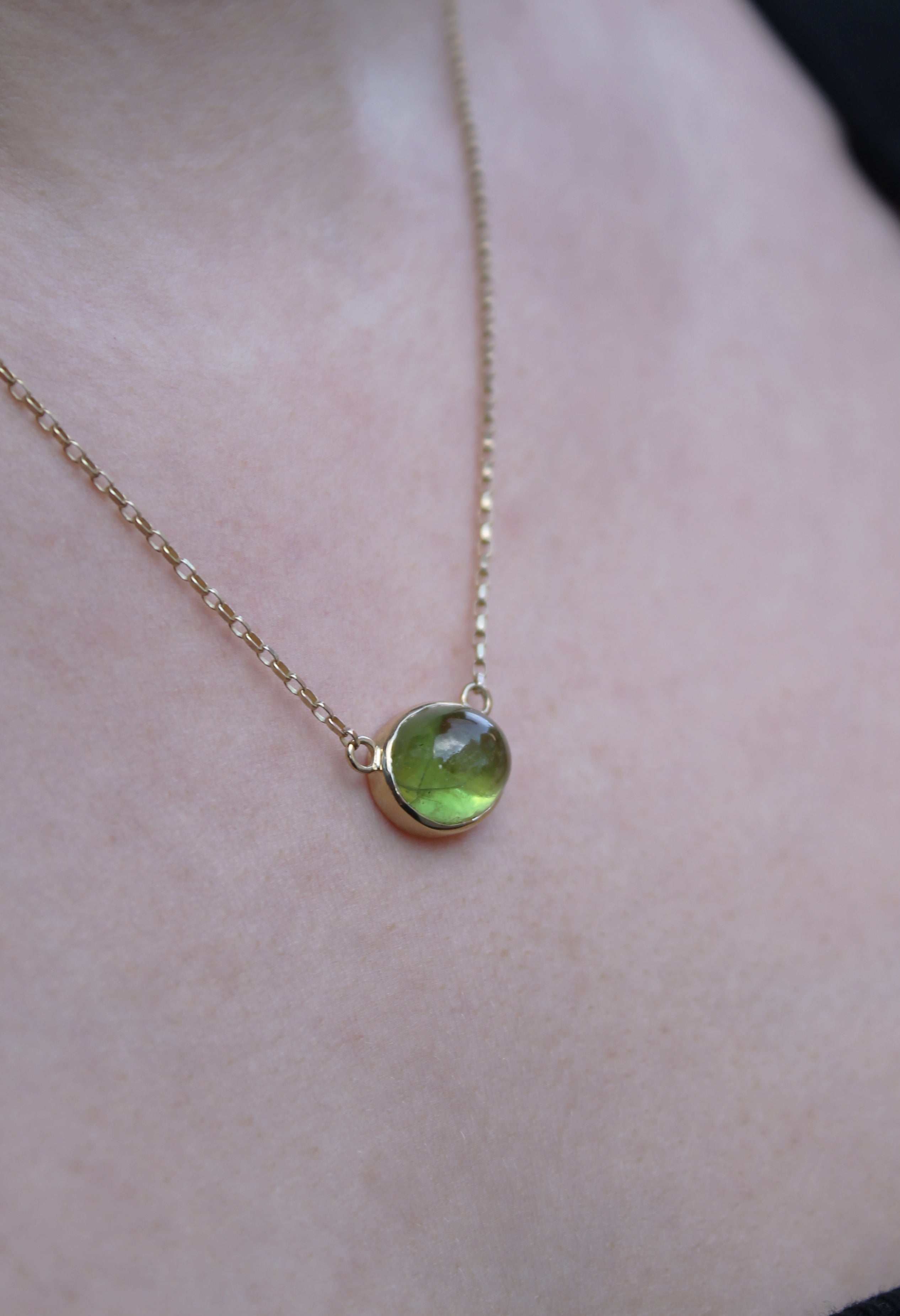 The Whispering Moss 9ct Yellow Gold Necklace by Madeleine Fine Jewellery, featuring a round green peridot cabochon pendant, rests against a person’s bare skin.