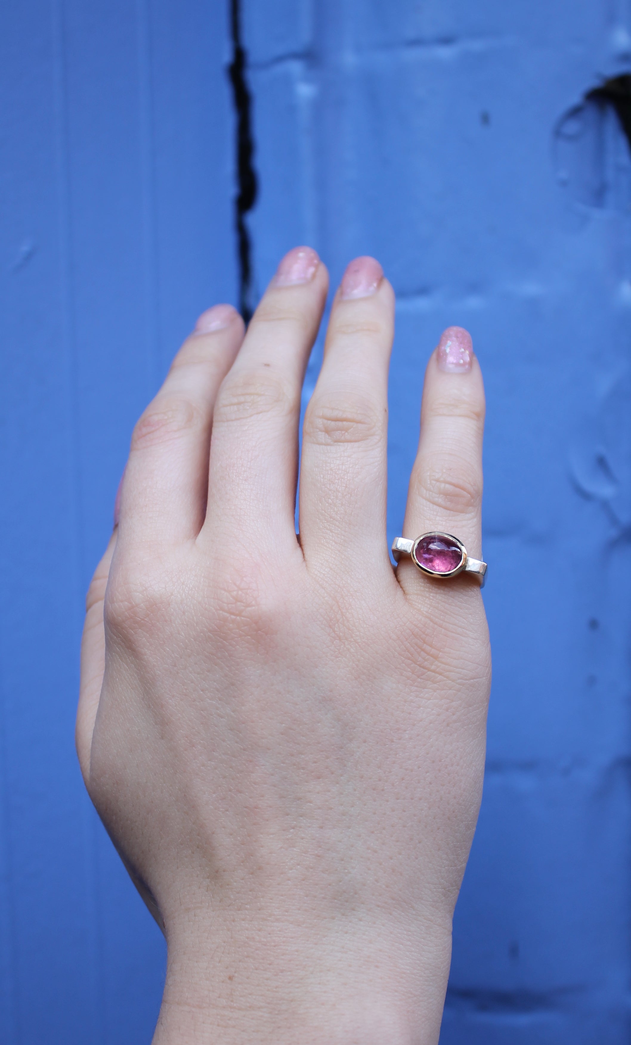 A hand with light skin and short, pink nails displays the Madeleine Fine Jewellery Stellar Blush Silver & Gold Ring, featuring an oval pink gemstone, against a textured blue background.