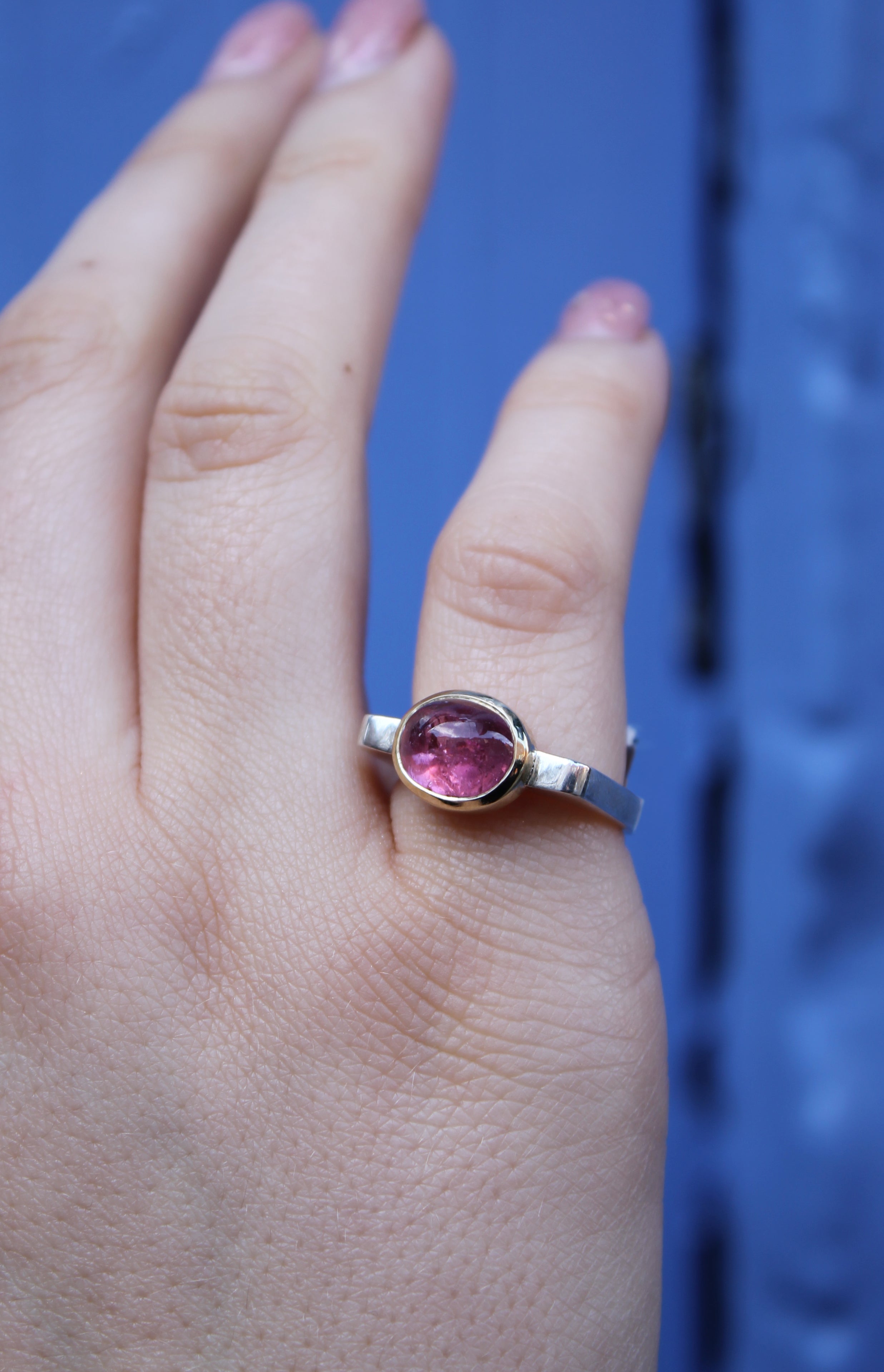 A hand with light skin and neat nails wears the Madeleine Fine Jewellery Stellar Blush Silver & Gold Ring, featuring a pink tourmaline oval cabochon, set against a blue background.