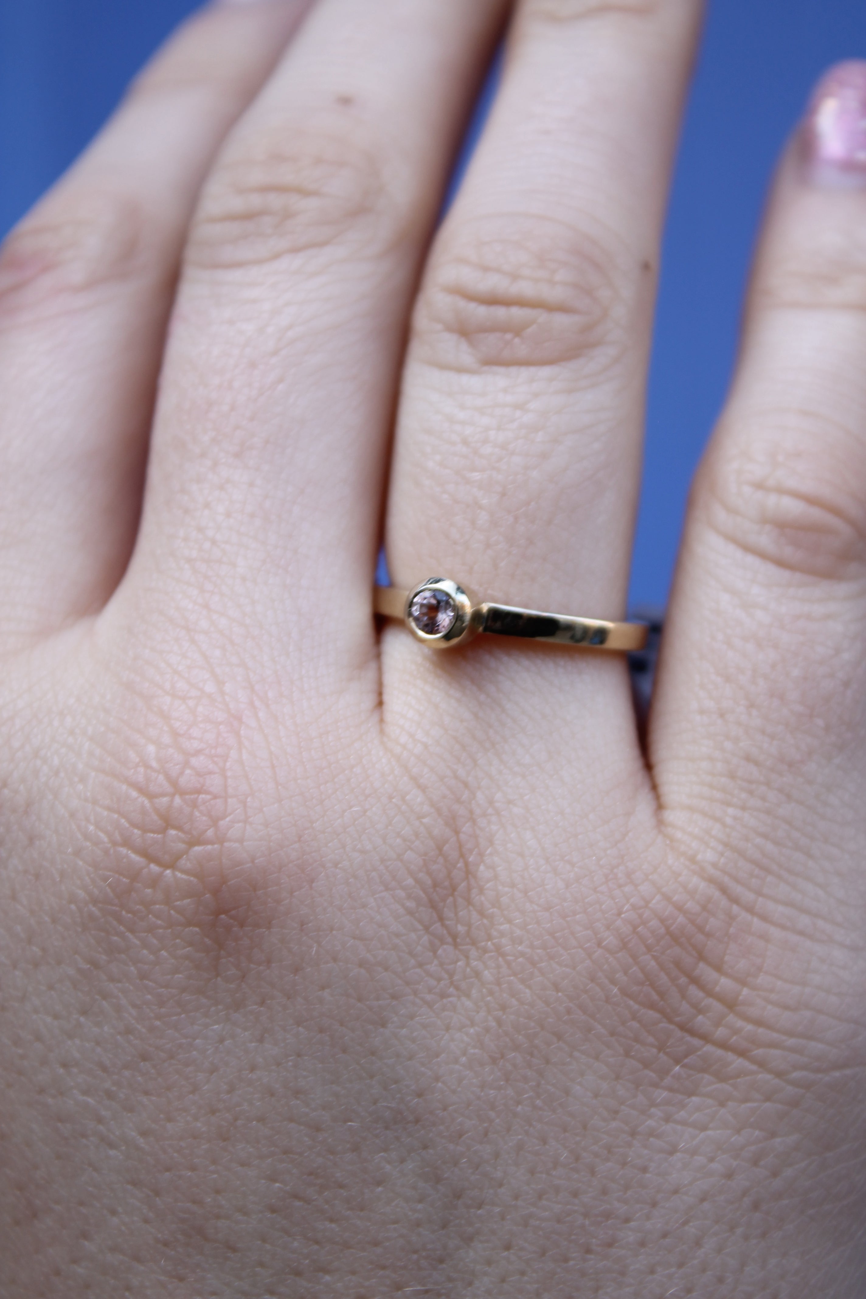 A close-up shows a hand with natural nails wearing the Madeleine Fine Jewellery Gilded Blossom 9ct yellow gold ring, featuring a small, round clear gemstone at its center, set against a blue background.