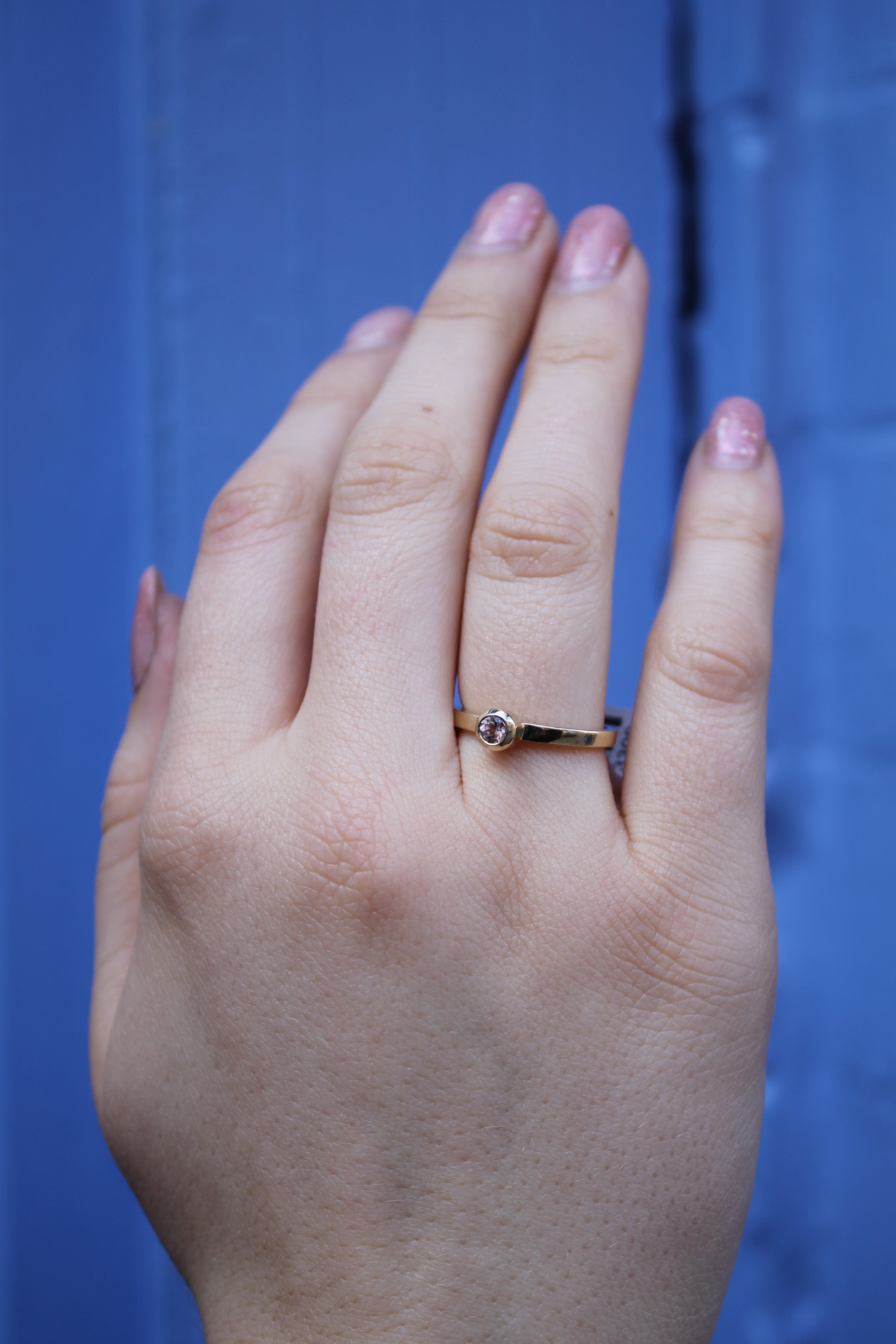A close-up of a light-skinned hand wearing the Madeleine Fine Jewellery Gilded Blossom 9ct yellow gold ring with a small clear stone on the ring finger, set against a blue background.